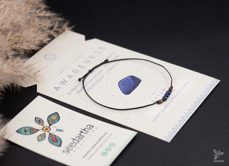 May include: A black string bracelet with blue lapis lazuli beads and a small lapis lazuli stone charm. The bracelet is on a card with the text "AWARENESS" and "WATER" on it.