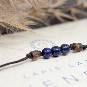 May include: A dark blue and brown beaded bracelet with a black cord. The beads are round and have a smooth, polished finish. The bracelet is lying on a white surface with the text "Lapis Lazuli" printed on it.