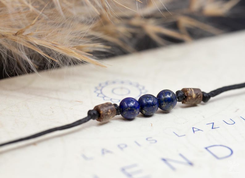 May include: A dark blue and brown beaded bracelet with a black cord. The beads are round and have a smooth, polished finish. The bracelet is lying on a white surface with the text "Lapis Lazuli" printed on it.