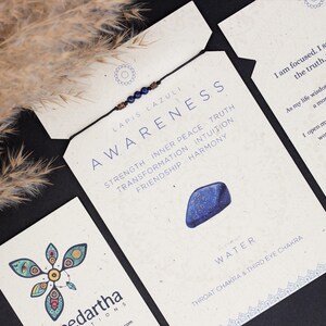 May include: A blue lapis lazuli bracelet with a brown cord and a small blue stone charm. The card reads "Lapis Lazuli Awareness" and lists the benefits of the stone, including strength, inner peace, truth, transformation, intuition, friendship, and harmony. The card also states "Water, Throat Chakra & Third Eye Chakra".