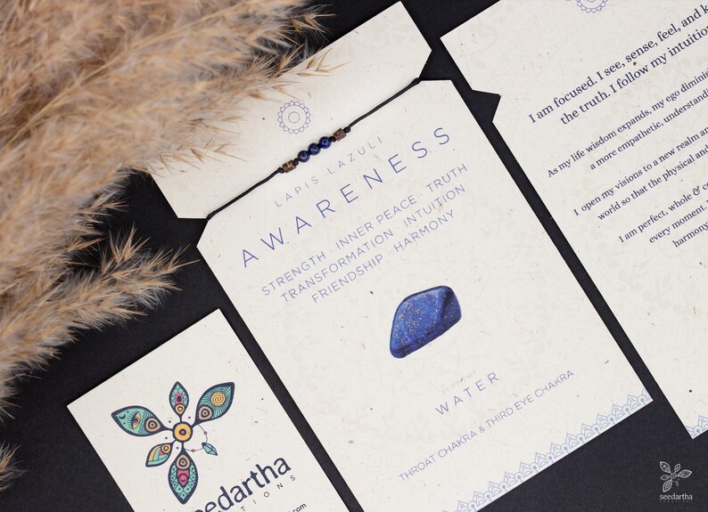 May include: A blue lapis lazuli bracelet with a brown cord and a small blue stone charm. The card reads "Lapis Lazuli Awareness" and lists the benefits of the stone, including strength, inner peace, truth, transformation, intuition, friendship, and harmony. The card also states "Water, Throat Chakra & Third Eye Chakra".
