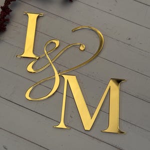 Könnte beinhalten: Goldfarbene, verspiegelte Acryl-Buchstaben 'I', 'J' und 'M' auf einer hellgrauen Holzoberfläche angeordnet. Das 'J' ist mit einem fließenden Schriftzug gestaltet, der mit dem 'M' verbunden ist. Ein Zweig mit bordeauxfarbenem Zierlaub im Hintergrund.