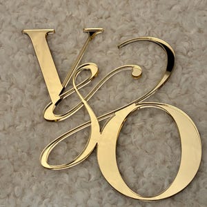 Könnte beinhalten: Goldfarbene Metallbuchstaben, die "VO" in einer dekorativen Schriftart bilden. Die Buchstaben sind reflektierend und liegen auf einer strukturierten, cremefarbenen Oberfläche. Das "V" ist aufrecht, das "O" rund.