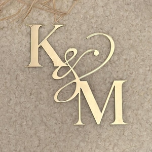 Könnte beinhalten: Ein goldfarbenes, reflektierendes Monogramm-Schild mit den Großbuchstaben K und M, die durch ein stilisiertes Et-Zeichen verbunden sind. Die Buchstaben sind in einer Serifenschrift mit glatter, polierter Oberfläche und Spiegeleffekt gehalten.