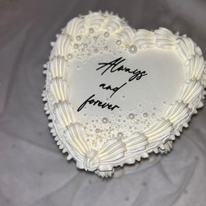 Könnte beinhalten: Ein herzförmiger Kuchen mit weißer Glasur, verziert mit einem gerüschten Rand und kleinen weißen essbaren Perlen. In der Mitte des Kuchens befindet sich der schwarze Schriftzug "Always and forever" in kursiver Schrift auf der glatten Oberfläche.