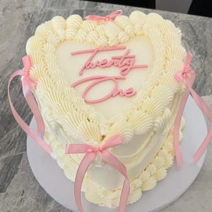 Könnte beinhalten: Ein herzförmiger Kuchen mit weißem Zuckerguss und rosa Schleifen. Der Kuchen ist mit den Worten "Twenty One" in rosa Schrift verziert. Der Kuchen steht auf einem weißen Teller.