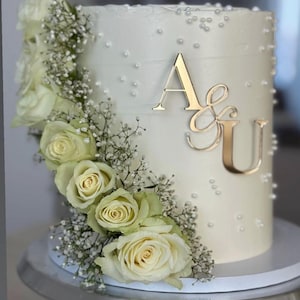 Könnte beinhalten: Eine weiß glasierte Torte, verziert mit weißen Rosen und Schleierkraut. Goldfarbene Initialen "A&U" zieren die Seite. Die Torte steht auf einem silbernen Tortenständer und hat kleine weiße Perlenspritzer.