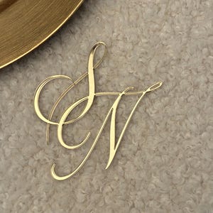 Könnte beinhalten: Ein goldfarbenes Acryl-Monogramm mit den ineinander verschlungenen Buchstaben S und N in einer Schreibschrift. Die Buchstaben haben eine glatte, reflektierende Oberfläche und sind zu einem Stück verbunden, mit eleganten, fließenden Linien.