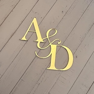 Könnte beinhalten: Goldfarbenes Monogramm mit den Buchstaben "A" und "D" in dekorativer Schrift. Die Buchstaben sind mit einem kaufmännischen Und verbunden. Das Monogramm wird auf einem hellgrauen Holzgrund angezeigt.