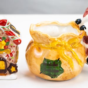 Può includere: Un set sale e pepe in ceramica a forma di casetta di pan di zenzero con tetto a righe rosse e bianche, e un contenitore in ceramica a forma di sacco regalo con una statuina di Babbo Natale, dipinti in rosso, verde, giallo e marrone.