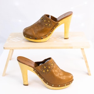 Pode incluir: Um par de tamancos de couro castanho com saltos e solas de madeira. Os tamancos têm tachas douradas e um salto de madeira clara. São exibidos numa pequena mesa de madeira. Os saltos têm cerca de 10 cm de altura.