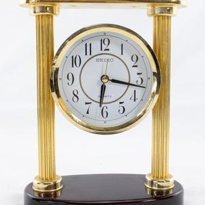 Puede incluir: Un reloj de escritorio de cuarzo Seiko dorado con esfera blanca y números negros. El reloj está sostenido por dos columnas estriadas y una barra superior rectangular, todo en dorado. La base es de madera oscura y pulida. Las manecillas del reloj son negras.