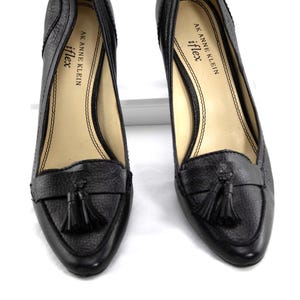 May include: A pair of black leather heeled loafers with a textured finish. Each shoe features a small tassel detail and the brand name "AK Anne Klein iflex" printed on the insole.