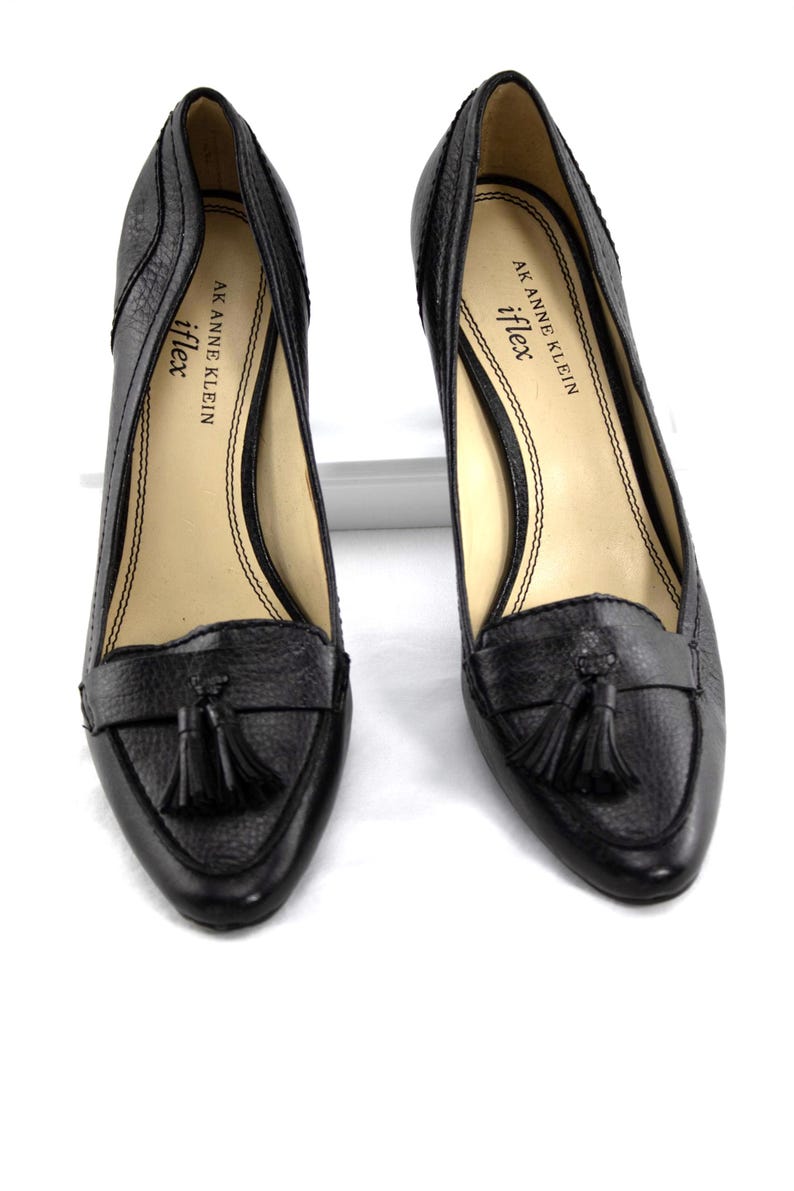 May include: A pair of black leather heeled loafers with a textured finish. Each shoe features a small tassel detail and the brand name "AK Anne Klein iflex" printed on the insole.