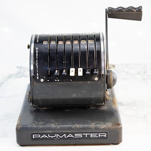 May include: A vintage black Paymaster cash register with a handle and number dials for dollars and cents. The register has a "PROTECTION" label on the side and the "PAYMASTER" logo on the base. The register is likely from the mid-20th century.