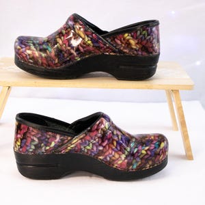 Könnte beinhalten: Clogs mit einem farbenfrohen, strickähnlichen Muster in Lila, Rosa, Gelb und Grün. Die Clogs haben eine schwarze Sohle und Zierleiste. Einer steht auf einem kleinen Holztisch, der andere darunter.