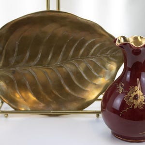 May include: A brass leaf-shaped tray resting on a gold metal stand, positioned behind a small, round, deep red ceramic vase with a ruffled gold-rimmed opening and a gold floral pattern painted on the front surface of the vase.