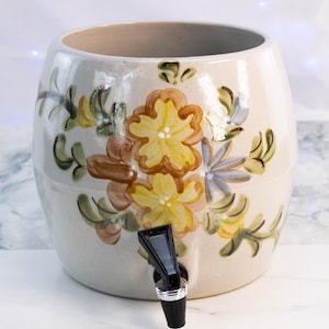 Puede incluir: Dispensador de bebidas de cerámica color crema con un diseño floral en amarillo, marrón y azul. El dispensador tiene un grifo negro y una amplia abertura en la parte superior. El patrón floral está pintado a mano.