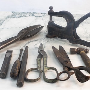May include: An assortment of antique metal tools, including large shears, chisels, scissors, a hammer, and a lever press. The tools are arranged on a white and gray marble surface, showcasing their aged, rustic appearance.