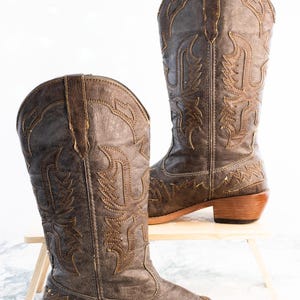 May include: Two brown cowboy boots with detailed stitching. The boots feature a pointed toe, a mid-height heel, and decorative stitching in a lighter brown. They are presented on a small wooden stand against a white backdrop.