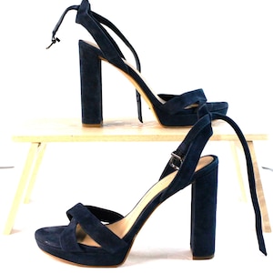 Könnte beinhalten: Ein Paar marineblaue Wildleder-Plateausandalen mit dicken Blockabsätzen. Die Sandalen haben Knöchelriemen und einen Schnallenverschluss. Sie werden auf einem kleinen, hellen Holztisch vor weißem Hintergrund präsentiert.