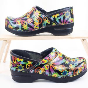 May include: A pair of black clogs featuring a vibrant, multicolored pattern of peace signs, hearts, and flowers. The shoes have a glossy finish, a rounded toe, and a thick, solid black sole with a slight heel, providing a sturdy and slip-on design.