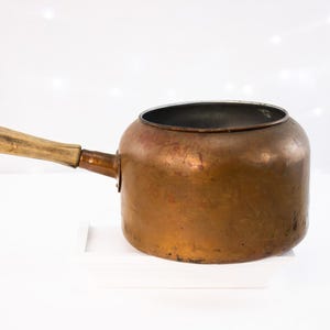 May include: A copper saucepan with a wooden handle. The pot has a rounded body and a handle attached with rivets. The handle is made of light-coloured wood. The saucepan is sitting on a white surface.