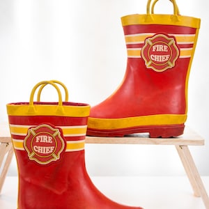 Könnte beinhalten: Ein Paar rote Gummistiefel mit gelben Rändern und Zugschlaufen. Jeder Stiefel hat zwei horizontale gelbe Streifen und ein kreisförmiges Emblem an der Seite mit rotem Rand und dem Text FIRE CHIEF in weißen Buchstaben auf rotem Grund.