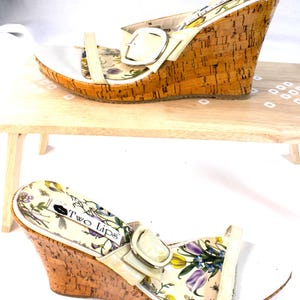 Puede incluir: Sandalias de cuña Two Lips con suela de corcho y correas blancas. Las sandalias tienen un estampado floral en la plantilla y un cierre de hebilla. El tacón de cuña mide aproximadamente 10 cm de alto.