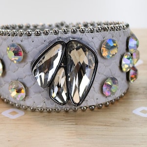 May include: Three bracelets: a thin gold-tone band with small clear crystals, a wide light gray textured cuff featuring large iridescent and clear faceted stones, and a dark metal link bracelet with rectangular multicolored glass inlays.