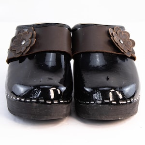 May include: Pair of black clogs with a glossy finish, featuring a brown leather strap and a decorative flower accent. The wooden soles are dark brown with white stitching around the edges. A stylish and comfortable footwear option.