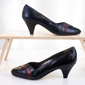 May include: A pair of black pumps with a textured snakeskin pattern. Each shoe features a rounded toe with a decorative band across the vamp in dark green and deep red. The footwear has a mid-height kitten heel and a smooth interior lining.