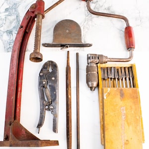May include: An assortment of vintage hand tools, including a red drill press, a brace and bit, a pair of pliers, files, and a drill bit set in a wooden box. The tools are arranged on a white surface.