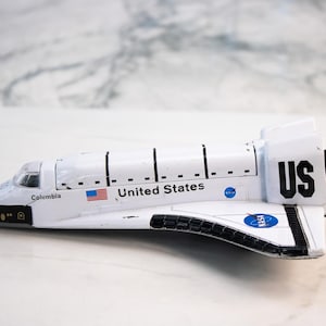 Puede incluir: Una maqueta blanca de transbordador espacial con detalles en negro, con las palabras "United States" y la bandera estadounidense. El transbordador tiene la palabra "Columbia" y las letras "US" en la cola. Modelo de vehículo espacial.