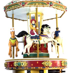 May include: A decorative carousel with three toy horses and riders. The carousel features a red and gold base, a white canopy with green and gold trim, and gold support poles. The horses are cream-colored with black manes and tails.