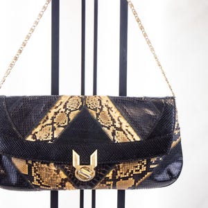 May include: A rectangular clutch bag featuring a snakeskin-patterned texture in black and tan. The bag has a fold-over flap with a central black triangular panel and a gold-tone metal clasp. It includes a thin gold-tone metal chain shoulder strap.