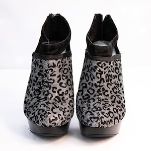May include: A pair of black and silver platform booties. The booties feature a black suede upper with a silver leopard print design on the front. The platform and heel are black. The booties have a zipper closure at the back.