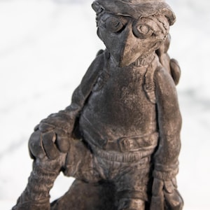 Puede incluir: Una escultura gris oscuro de una criatura con forma de pájaro que lleva sombrero, mochila y botas. La figura está posada con una pierna doblada, apoyada sobre una base de piedra. La escultura tiene una superficie texturizada y detalles.
