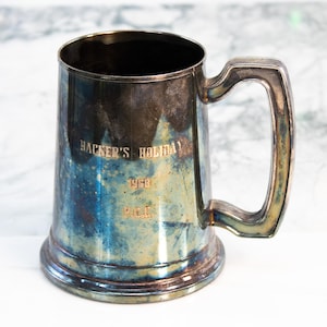 May include: A vintage silver beer mug with a handle. The mug has a tarnished appearance with blue and silver hues. The text "HACKER'S HOLIDAY 1968 P.E.C." is engraved on the front. The mug sits on a white surface.