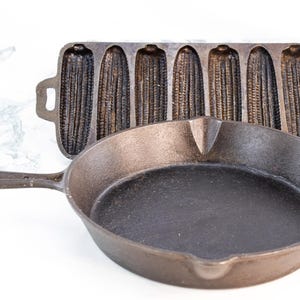 May include: A cast iron frying pan with a long handle and a corn-shaped cornbread mould. The frying pan is round and black, and the cornbread mould has six corn cob impressions. Both are made of dark metal.