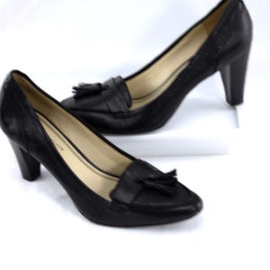 May include: A pair of black leather heeled loafers with a textured finish. Each shoe features a small tassel detail on the vamp and a block heel. The shoes are displayed on a white surface.