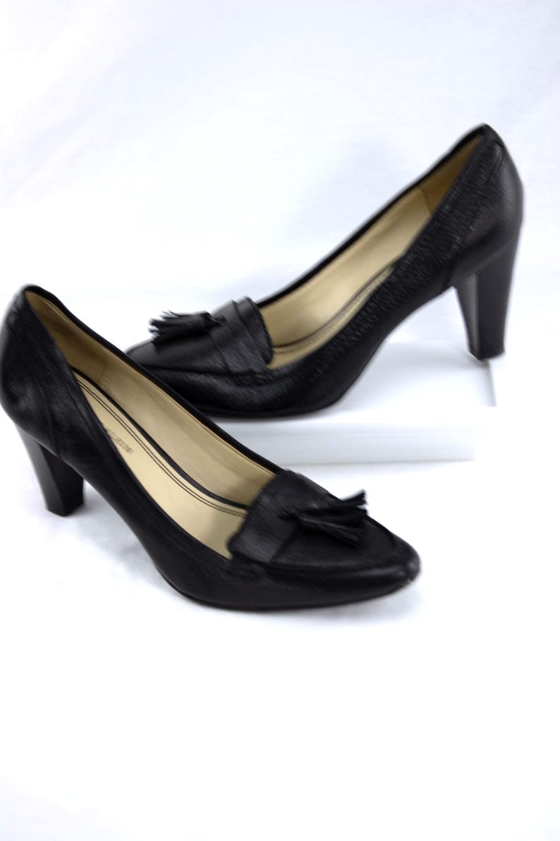 May include: A pair of black leather heeled loafers with a textured finish. Each shoe features a small tassel detail on the vamp and a block heel. The shoes are displayed on a white surface.