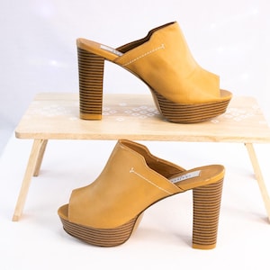 May include: Two pairs of open-toe platform heels in a light tan color. The heels have a thick, striped design, and the platforms are also striped. The shoes are displayed on a small wooden table with a white background.