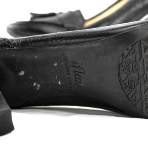 May include: Close-up of a black leather shoe with a block heel. The sole features a geometric tread pattern and the brand name "Flex". The shoe has a small bow detail on the top.
