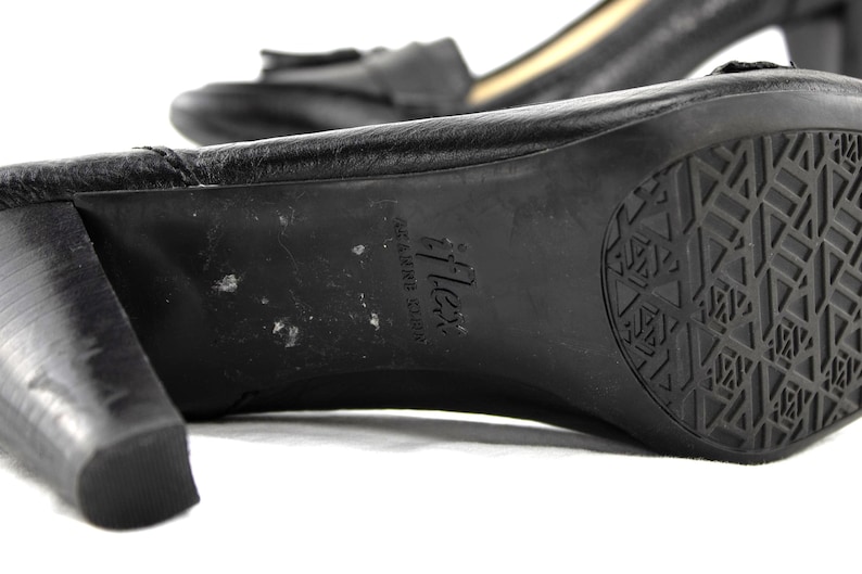 May include: Close-up of a black leather shoe with a block heel. The sole features a geometric tread pattern and the brand name "Flex". The shoe has a small bow detail on the top.