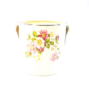 Puede incluir: Cubeta de hielo de cerámica blanca con un diseño floral con flores rosas y blancas, hojas verdes y un borde dorado. La cubeta tiene dos asas plateadas.