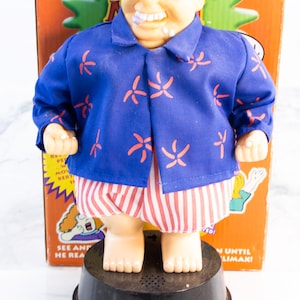 May include: A novelty toy figure of a bald man wearing glasses, a blue shirt with orange floral patterns, and red and white striped shorts. The figure is mounted on a black base, with a box in the background.