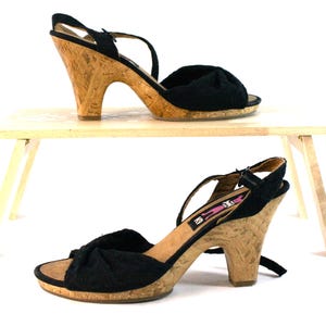 Puede incluir: Dos pares de sandalias negras de punta abierta con tacones y plataformas de corcho. Las sandalias tienen un detalle de tela anudada en la tira del dedo y correas de tobillo ajustables. Los tacones miden unos 10 cm de alto.