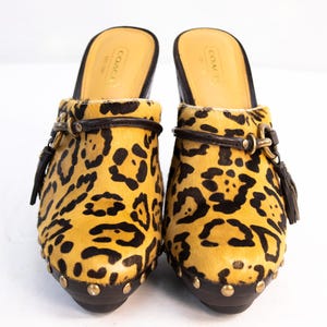 May include: A pair of Coach clogs with a leopard print design. The clogs have a yellow and black pattern, brown leather accents, and gold-tone hardware. The soles are dark brown with gold studs. The interior is tan leather with the Coach logo.