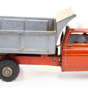 Pode incluir: Um caminhão de brinquedo basculante vintage com cabine laranja brilhante, detalhes de listras brancas e uma caçamba de metal cinza desgastada. O veículo possui pneus de borracha preta e um chassi vermelho, apresentando sinais de uso.